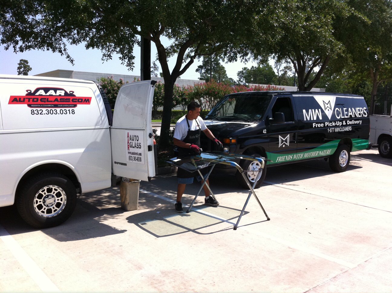 A person handling glass on a stand beside an AAR Auto Glass service van and an MW Cleaners van.